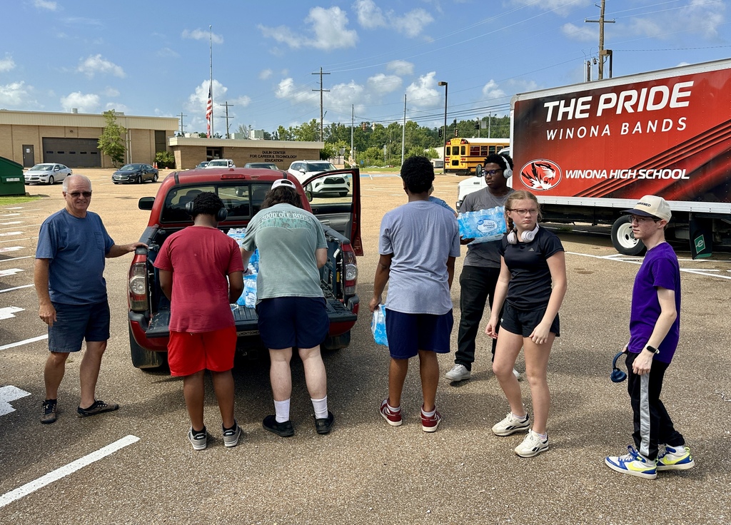 People load bottled water into a pickup truck. They stand on a parking lot with a red truck in the background.
