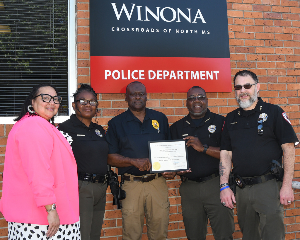 Four people stand in front of a brick building, smiling. One holds a certificate. A sign above reads "Winona Police Department".