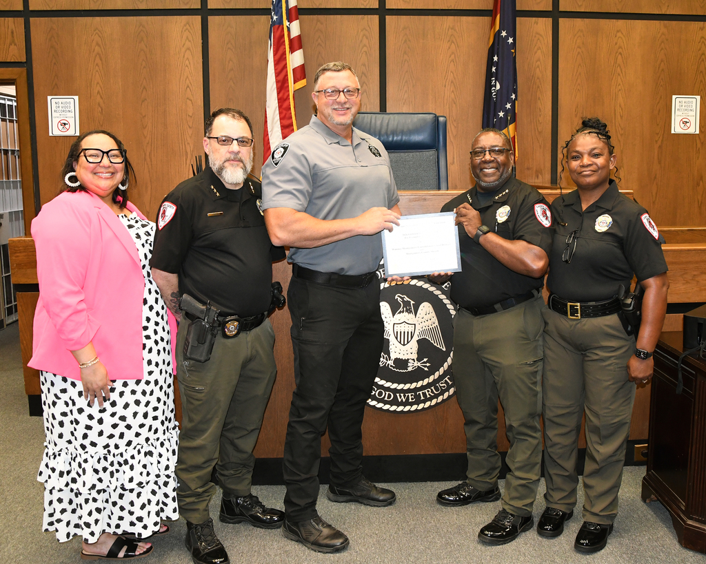 Five adults in uniform stand with a person in a pink dress in front of a wooden desk.
