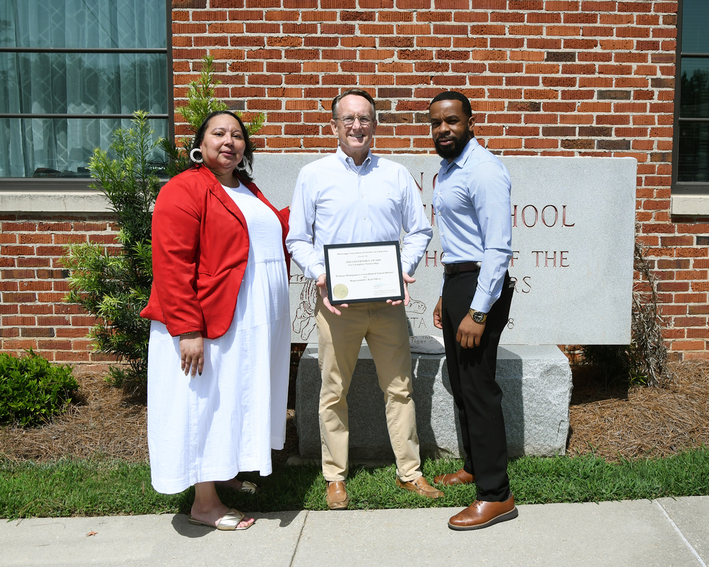Three people stand outside with a brick building behind them. A woman and two men hold a plaque.