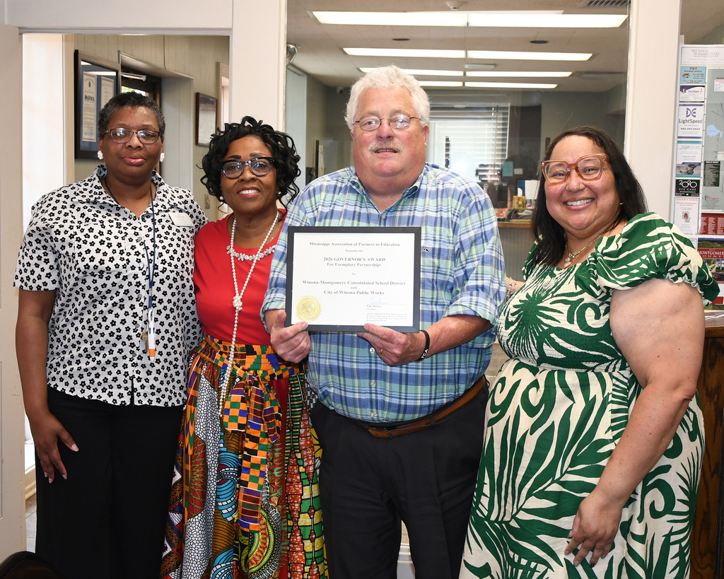Four adults stand close together, smiling. A man holds a framed certificate, while a woman stands next to him.