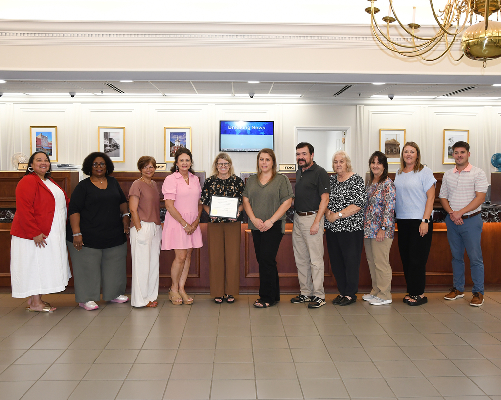 Nine individuals stand in a group with one holding an award. They are in a room with tiled floors, a chandelier, and framed pictures on the wall.