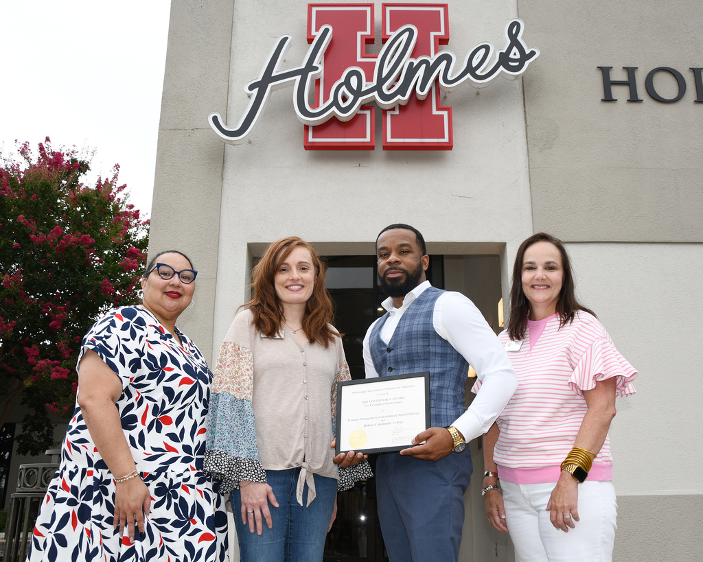 Four adults stand in front of a building, one man holds a framed certificate. A sign reads "Holmes" with red letters.