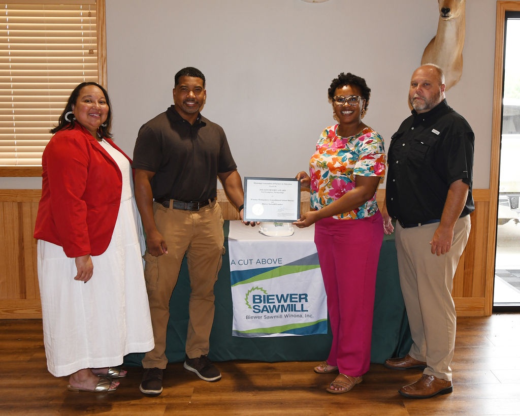 Four people stand in front of a banner, one holding a certificate. They are in a room with wooden floors.