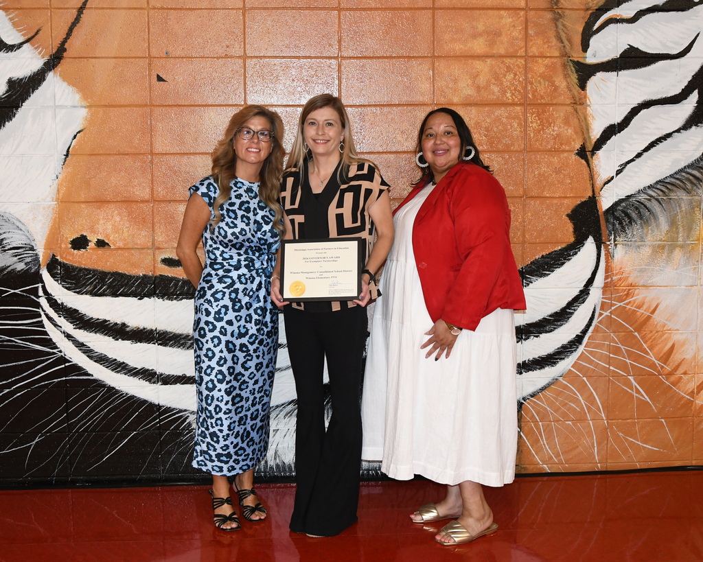 Three women in different outfits stand in front of a large tiger mural, one holding an award.