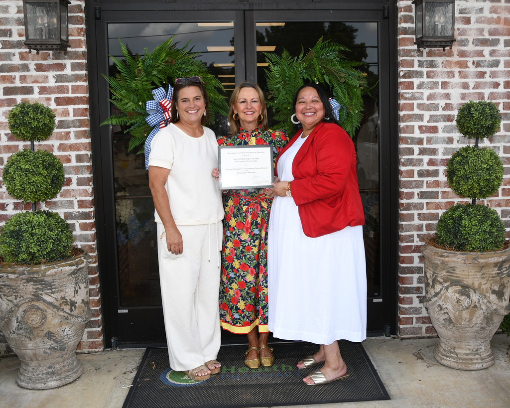 Three women stand in front of a brick building, one holding a framed certificate. Each woman wears summer clothing and sandals.
