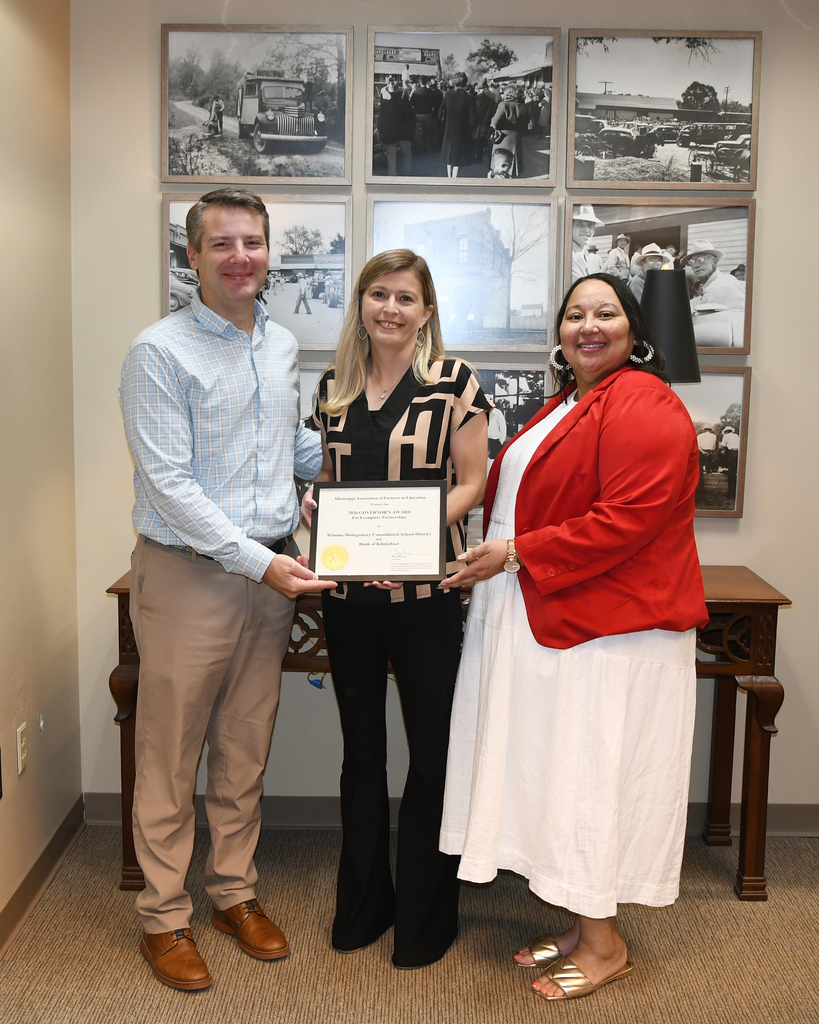 Three people hold a framed award. A man and a woman stand on either side of a woman. A desk with a lamp sits behind them.