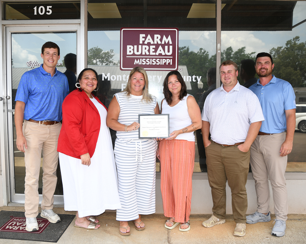 Six people pose for a picture in front of a building labeled "Farm Bureau." They hold an award.