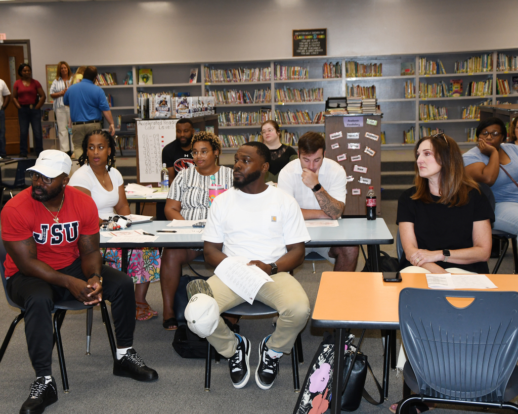A group of people are seated around tables in a library, looking at papers. One person wears a hat.