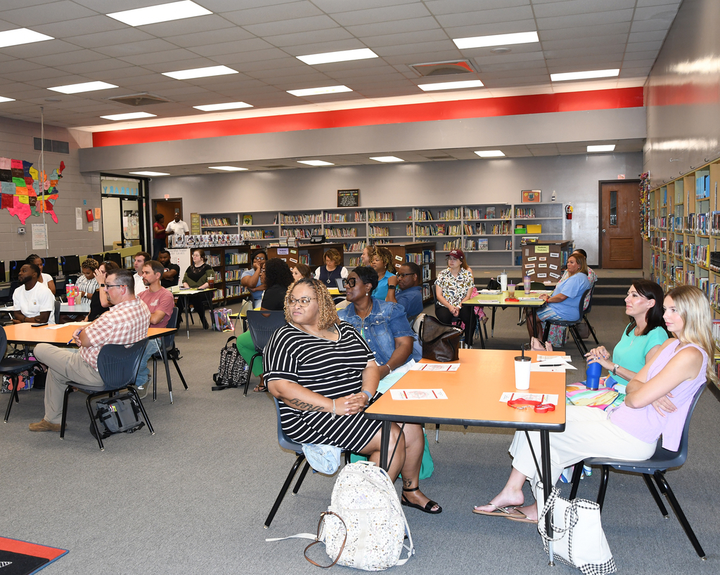 A group of people sit at tables in a library. Some wear casual attire and carry bags. Tables have items like books and cups.