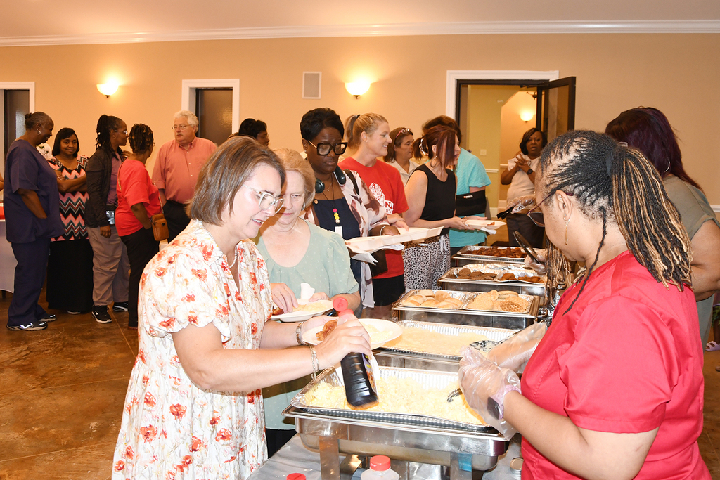 People gather in a room, with two women at a buffet table. A few trays of food are present.