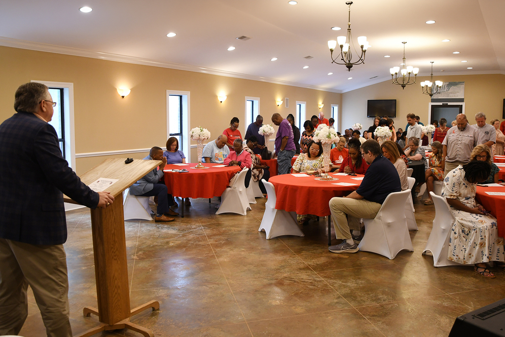 A man at a podium in a room with several tables with red tablecloths, chairs, and people.