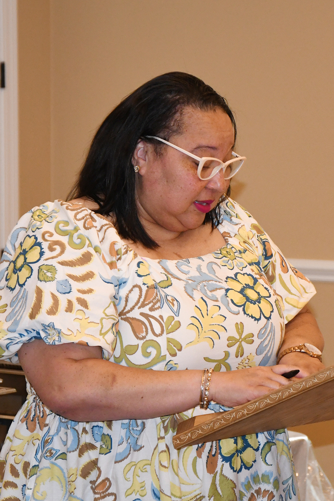 Woman in floral dress, glasses, and bracelets speaks into a microphone at a podium.