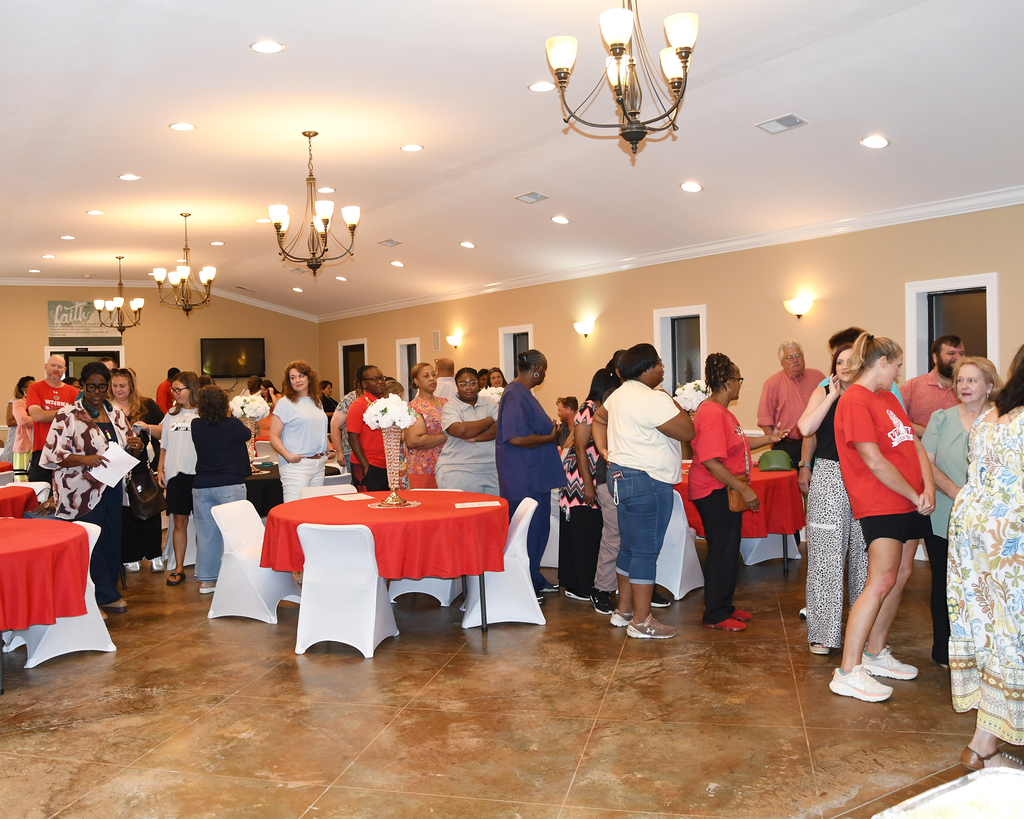Several people stand around tables in a room with a brown floor, white walls, and a chandelier.