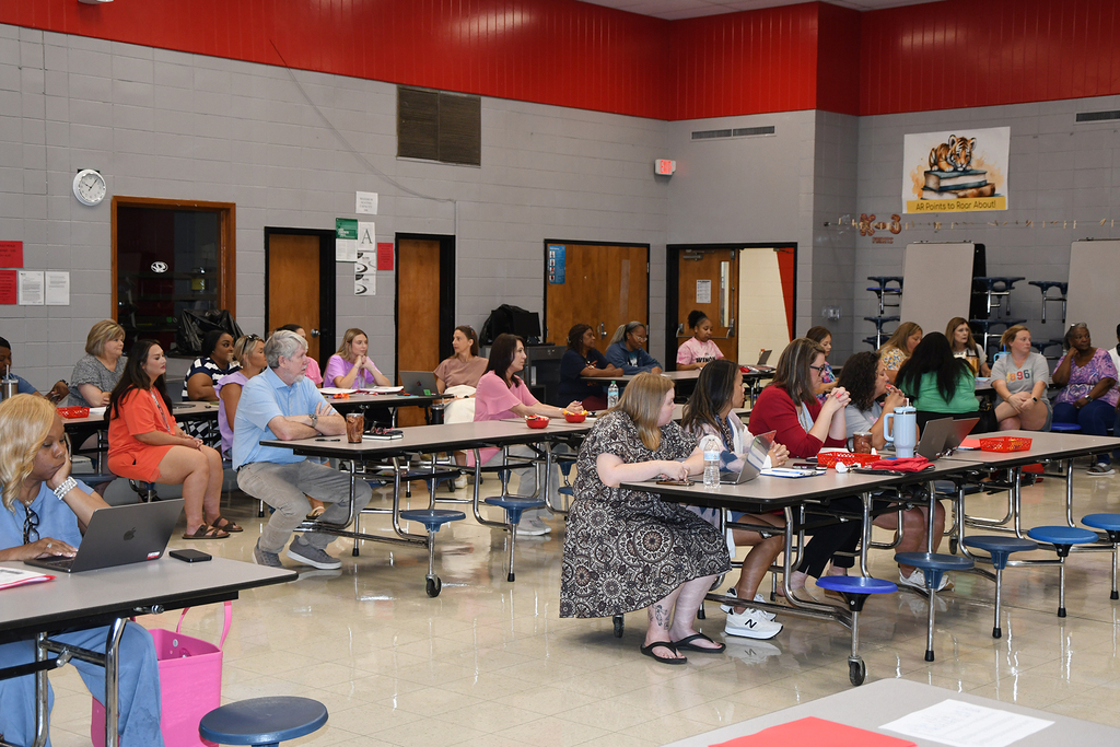 A cafeteria with people seated at tables. Tables have laptops, water bottles, and papers. Walls are gray with red trim.
