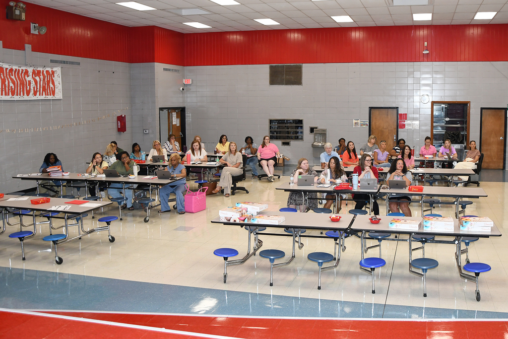 A classroom with tables and stools. Students sit at tables, some using laptops. Banners and doors in the background.