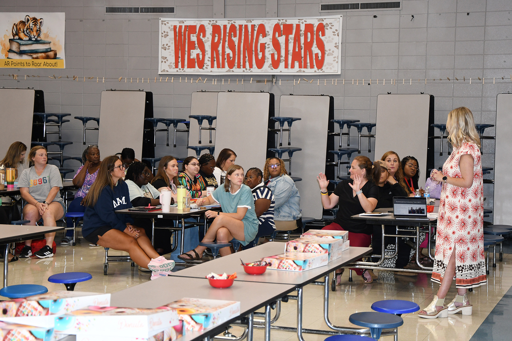A room with a banner reading "WES RISING STARS." Several individuals sit at tables; a woman stands at the front.