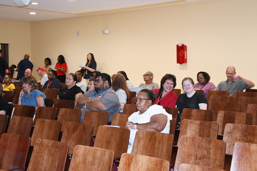 A conference room with rows of chairs. Several people sit facing the front, while others stand and speak at the back.