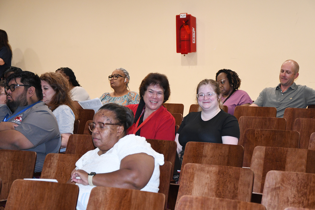 A group of adults seated in chairs in a room. Some wear glasses, watches, and bracelets. A fire alarm is on the wall.