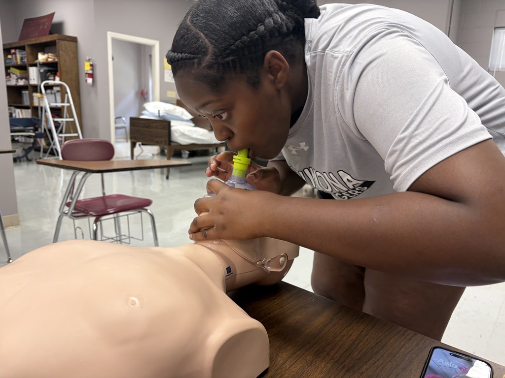 A woman in a white shirt performs CPR on a dummy in a room with chairs and a desk.