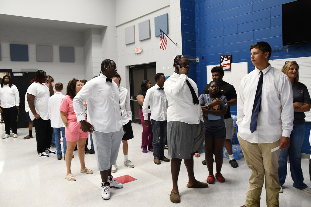 People in light-colored attire stand in a room with white and blue walls, some conversing. An American flag hangs on a wall, and a TV is mounted.