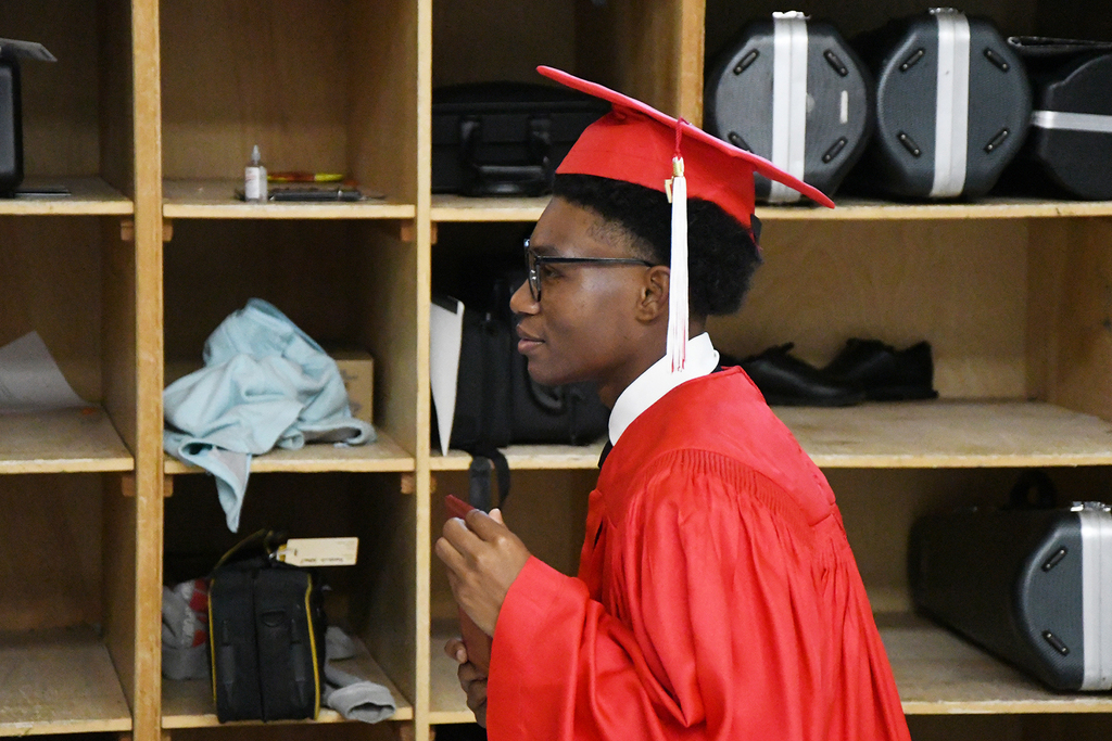 A young person in a red graduation gown and cap stands in a room, looking at something.