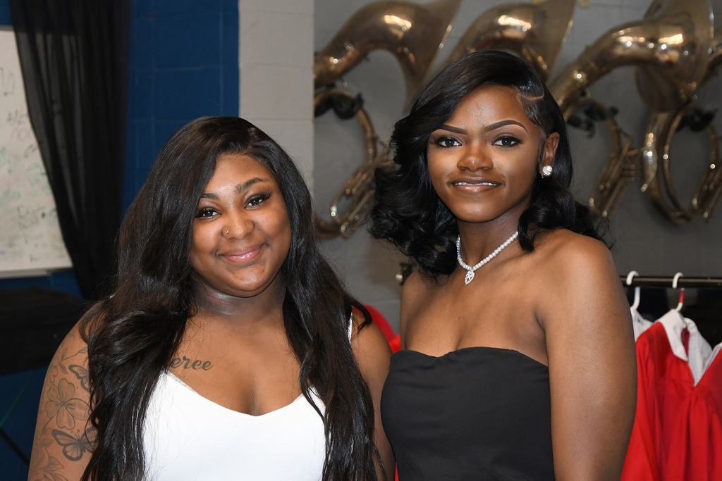 Two women smiling, one with long hair in a white dress, the other with short hair in a black dress, standing near a rack of red dresses.