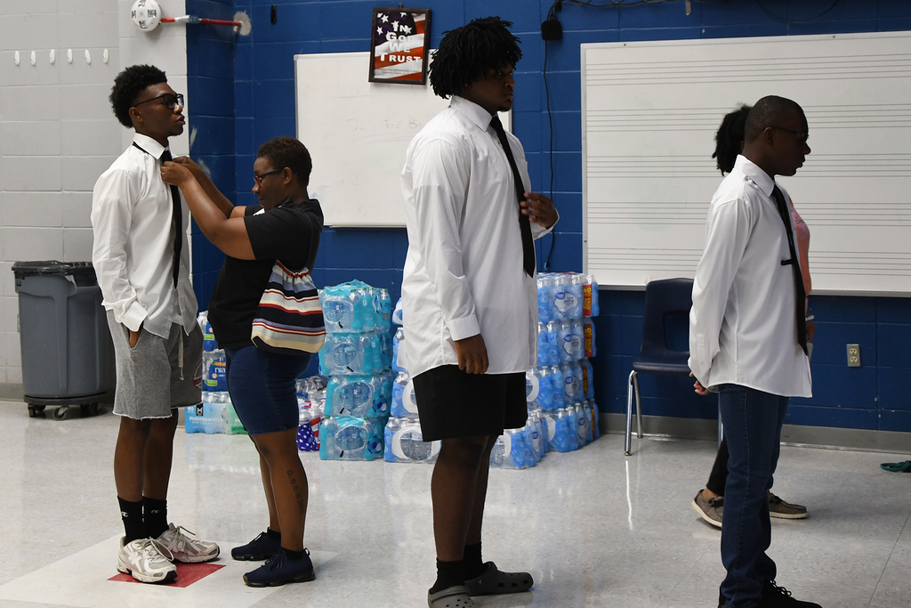 Four people in a room, three are adjusting ties on white shirts, one holding a bag. Blue wall with boards and a chair.