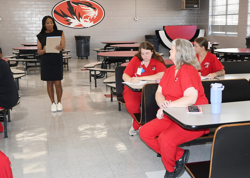 Several women in red scrubs seated at tables in a room. A woman in a black dress stands and holds a folder.