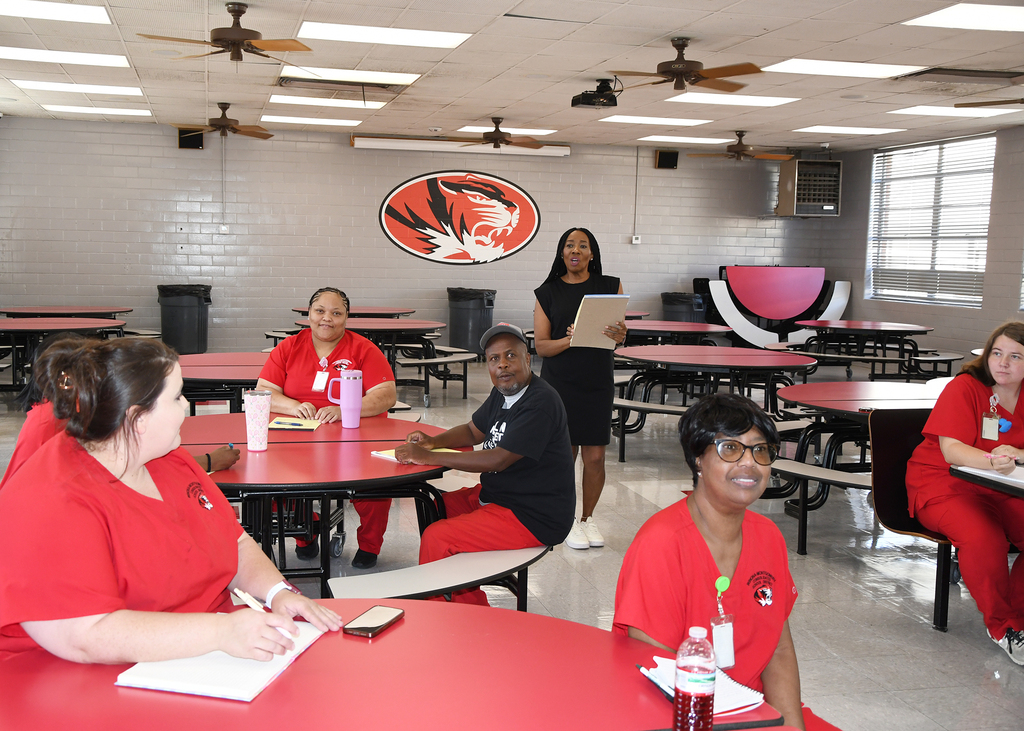 Staff in red uniforms seated around tables, one person holding a clipboard, one standing, and ceiling fans in a cafeteria.