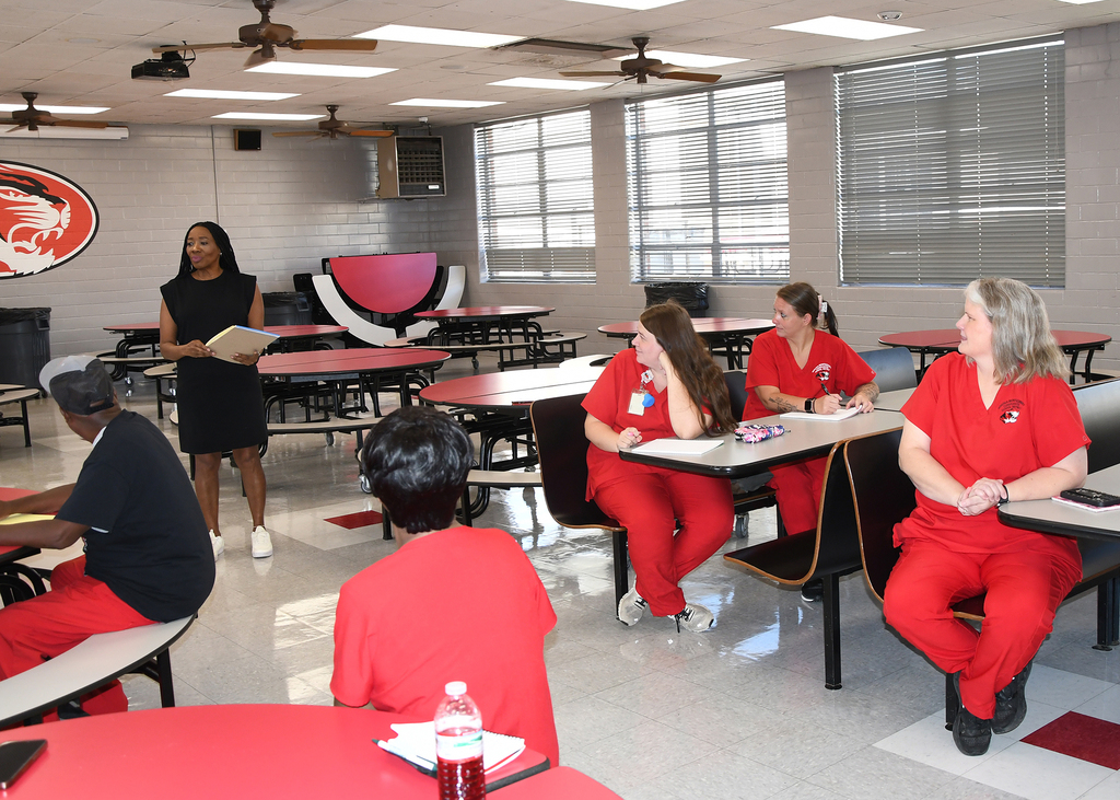People in red uniforms seated around tables in a room with a woman in black standing in front of them.