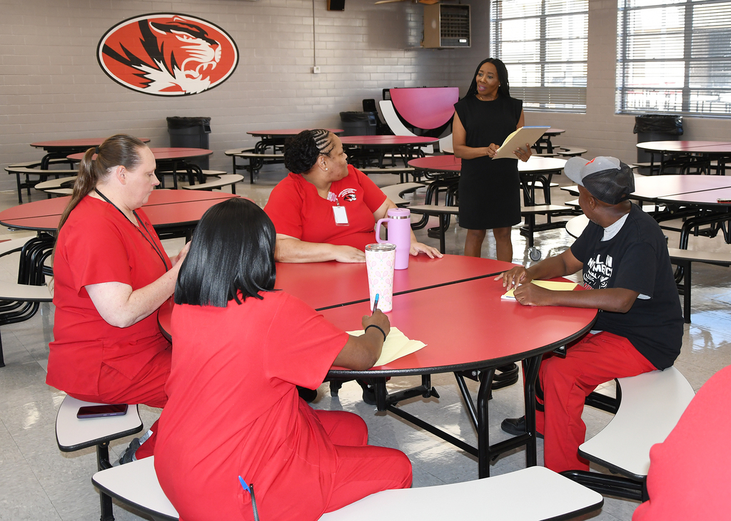 Six people sit around a table, with four wearing red uniforms. A woman in black stands behind them.