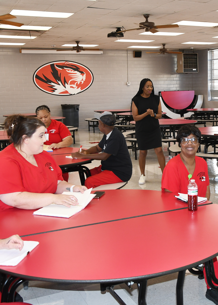 Four people sit around a table with papers and a bottle of water. A woman in a black dress walks near them. The room has red tables, a large tiger logo, and ceiling fans.
