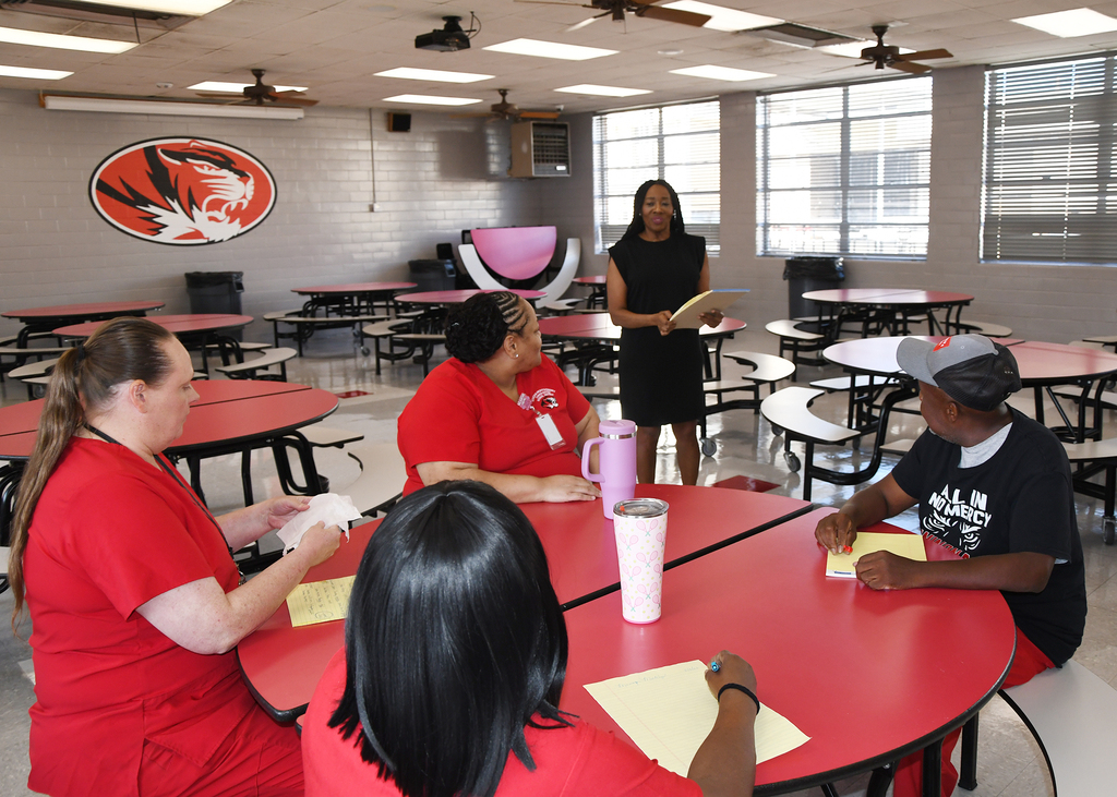 Four adults in red uniforms and one in black sit around a table. One adult holds a book.