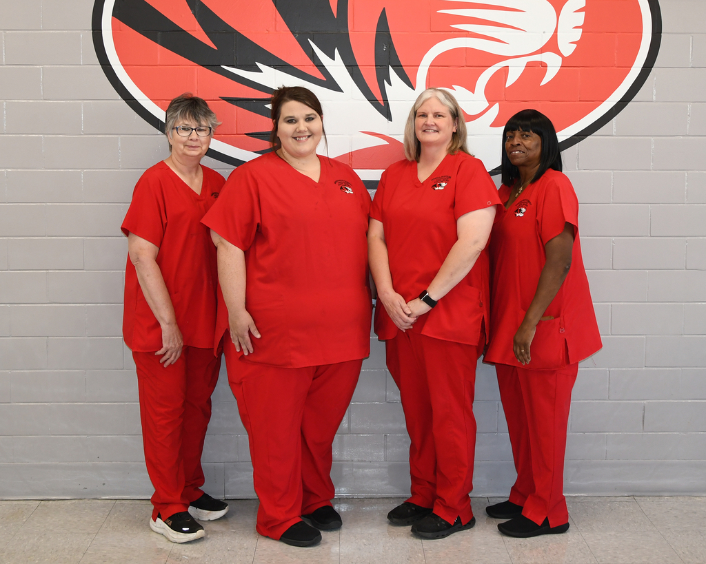 Five women in red scrubs stand next to each other in front of a white brick wall with a red logo.