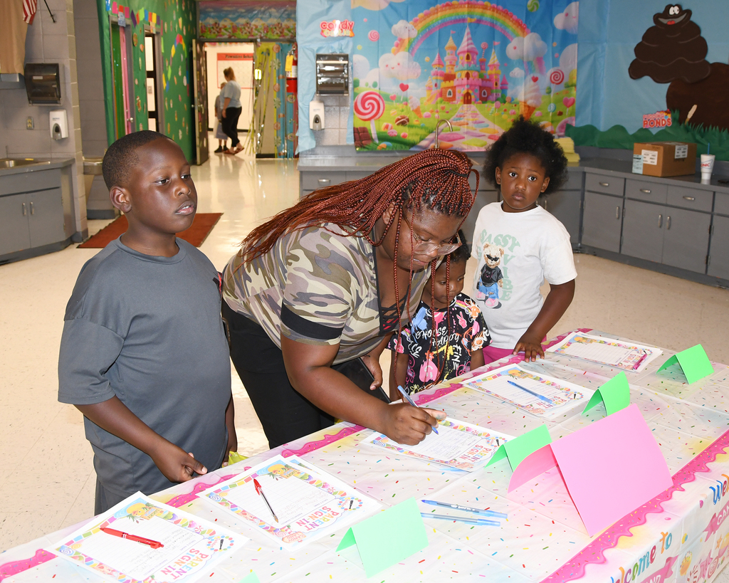 A woman leans over a long table, signing papers while two children stand nearby. Behind them, a room has colorful murals and cabinets.