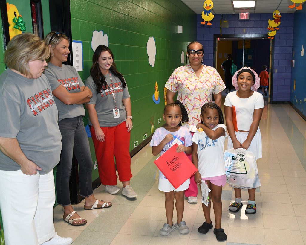 Group of adults and children in a hallway with green and blue walls. One child holds a red paper bag.
