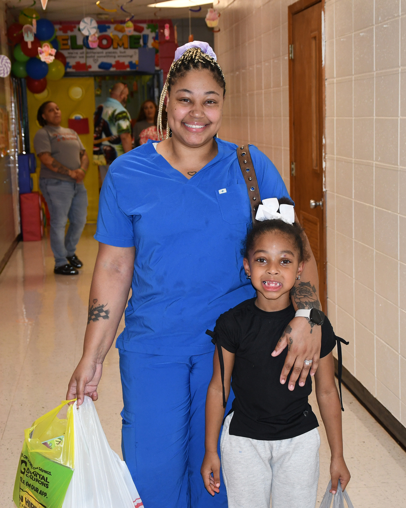 A woman in blue scrubs and a girl in black and white clothes walk in a school corridor, with bags in their hands.