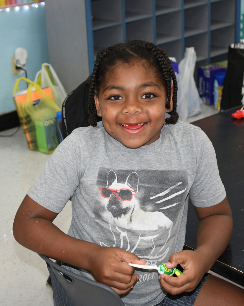 A child with braided hair in a gray tee sits at a table, smiling. A dog with sunglasses on the tee.