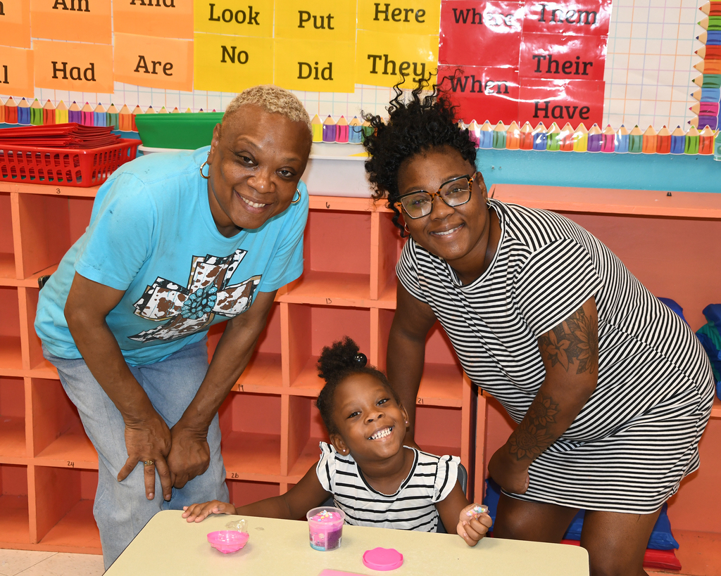 Two women and a young girl smile at camera in a classroom. Background includes shelves and a bulletin board.