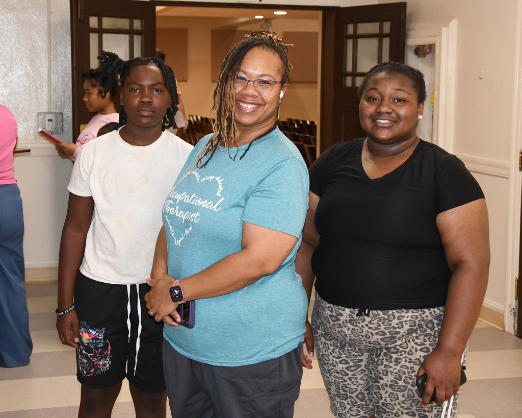 Three adults stand together, smiling at the camera. A woman in a blue shirt is in the center.