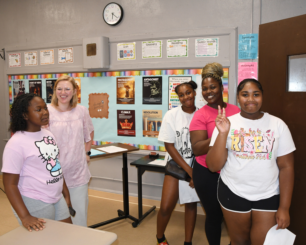 A group of four women stands in a classroom. One makes a peace sign. Two tables are behind them.
