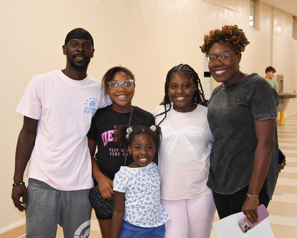 Five people standing in a hallway, smiling. A man in a pink shirt, two women in glasses, a girl in a floral shirt, and a young girl.
