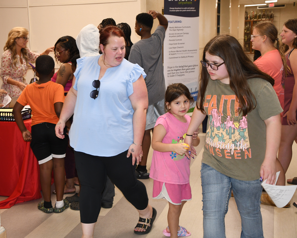 A woman and a young girl with glasses stand near a booth in a hallway. Behind them, a group of people is present.