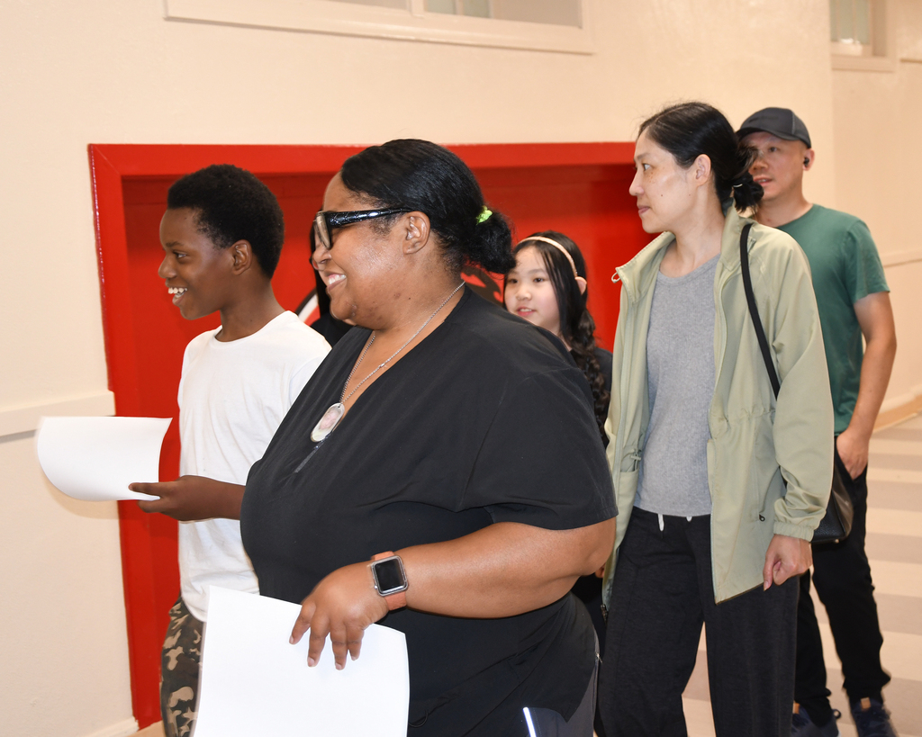 Five people stand in a room, one holding papers, with a red wall and a person in a green shirt in the background.