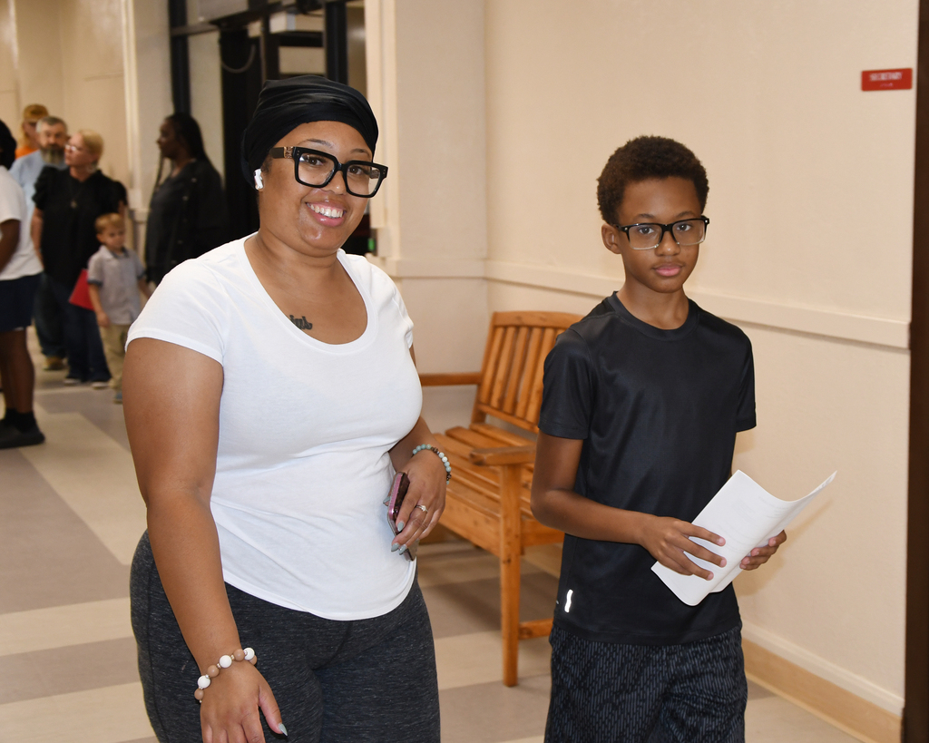 A woman in a white shirt and a boy in black stand in a hallway, holding papers. Behind them, a bench and people are visible.