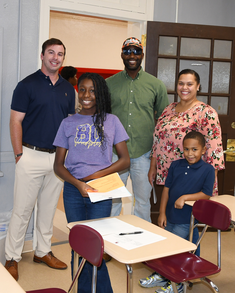 A group of four people stand in a classroom. A girl in a purple shirt holds a paper. A boy in a blue shirt stands near her.