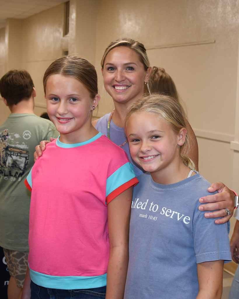 Two girls, one in pink, one in blue, smiling for a photo with a woman. Background shows a hallway with white walls and a ceiling.