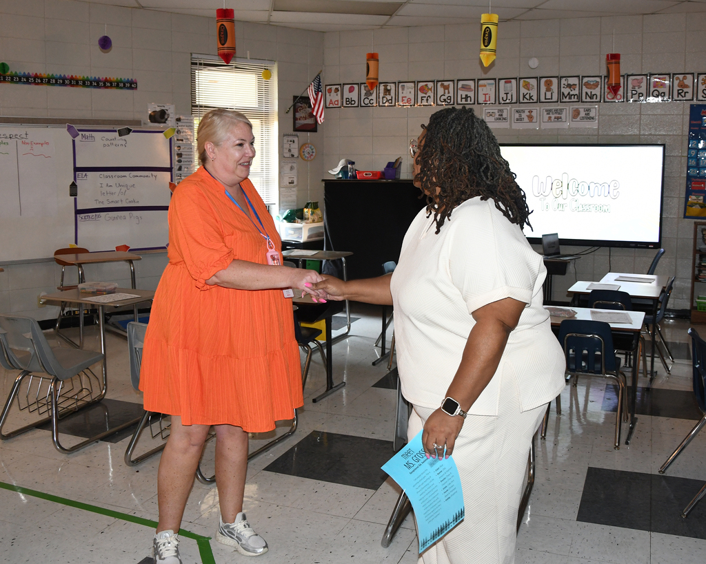 Two women shaking hands in a classroom. One wears an orange dress, the other white. Behind them, a whiteboard and projector screen.