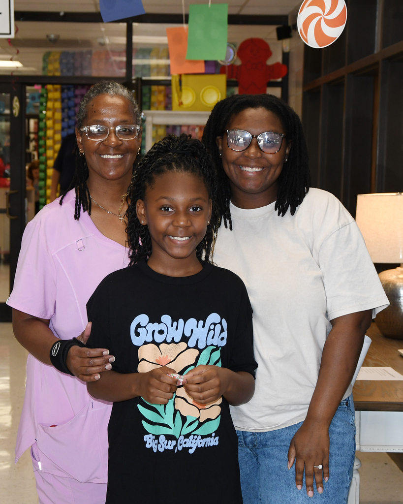 Three people standing in a room. A girl in a black t-shirt, a woman in pink, and a woman in white.